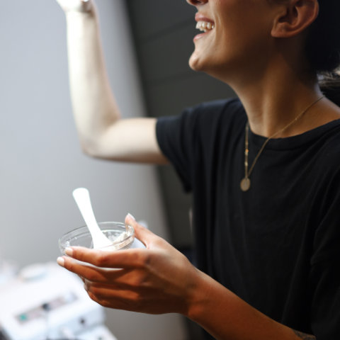 A woman in a black t-shirt laughs joyfully while holding a bowl and spoon near a window.
