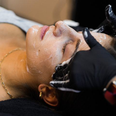 A woman receiving a facial treatment with a mask being applied to her face by a professional using black gloves.