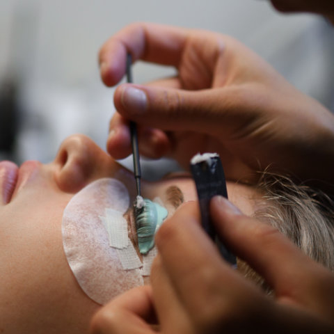 A close-up of a person receiving eyelash extensions with tweezers, showing their eyes covered with protective pads.