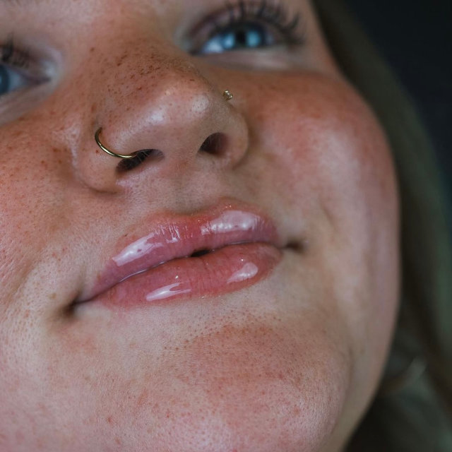 Close-up of a woman's lower face showing her nose and mouth, featuring freckles and a nose ring. she has glossy lips and is slightly smiling.