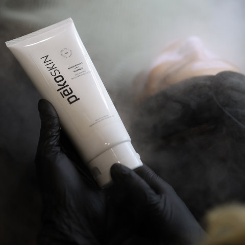 A hand in a black glove holding a tube of ren clean skincare body product, with a blurred figure in the background.